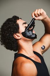 Close-up portrait of a man in a black tank top peeling a glossy black peel-off face mask from his cheek and nose, curly hair and small round sticker on his upper arm visible — men's skincare routine.