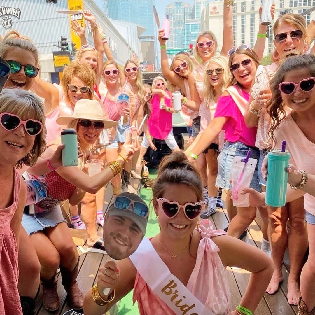 Fun bachelorette party on a sunny downtown outdoor bar patio — group of women in pink and heart-shaped sunglasses cheering with drinks, bride wearing a 'Bride' sash and holding a face cutout, city buildings in the background.