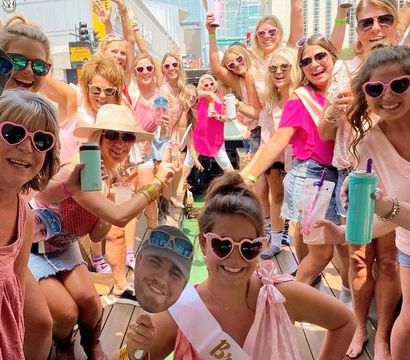 Fun bachelorette party on a sunny downtown outdoor bar patio — group of women in pink and heart-shaped sunglasses cheering with drinks, bride wearing a 'Bride' sash and holding a face cutout, city buildings in the background.