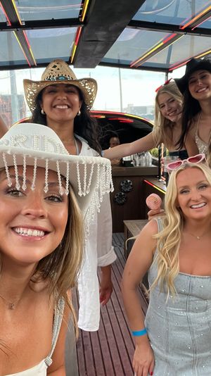 Smiling group of women on a neon-lit party trolley in the city, wearing cowgirl hats and summer dresses, holding drinks for a bachelorette-style outing