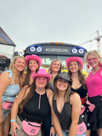 Seven smiling women wearing pink cowboy hats and matching pink fanny packs pose in front of a decorated party bus at an outdoor music festival