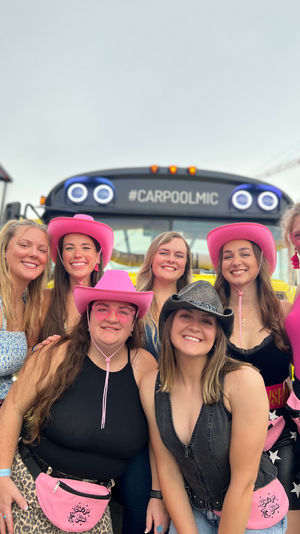 Seven women smiling in pink cowgirl hats and matching pink fanny packs posing in front of a colorful party bus at an outdoor music festival