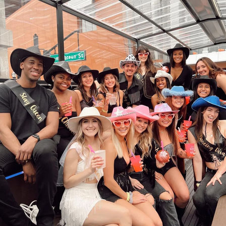 Cowgirl-themed bachelorette party on a covered rooftop patio — a large group wearing cowboy hats and sashes, smiling and holding colorful drinks in a downtown setting.