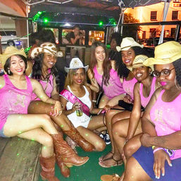Group of women in matching pink tank tops and cowgirl hats smiling inside a lit party bus at night, one wearing a sash and holding a drink — bachelorette celebration vibe.
