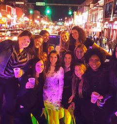 Smiling group of women celebrating on a neon-lit downtown nightlife street at night, holding drinks amid bustling traffic, bright storefronts and crowds in the background