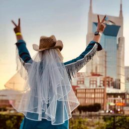 Person in cowboy hat, bridal veil and fringe jacket flashing peace signs in front of the Nashville skyline at sunset