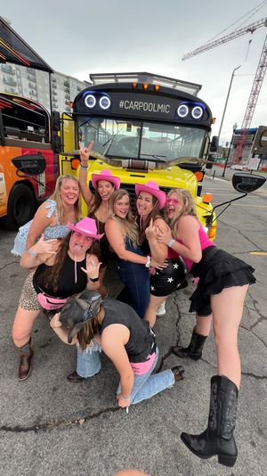 Group of friends in pink cowboy hats and party outfits laughing and posing in front of a bright yellow party bus labeled "#CARPOOLMIC" in a downtown parking lot with another colorful bus and a construction crane in the background.