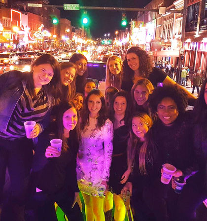 Group of smiling women on a neon-lit downtown bar street at night, posing for a night-out photo with drinks, traffic and bright signs in the background.