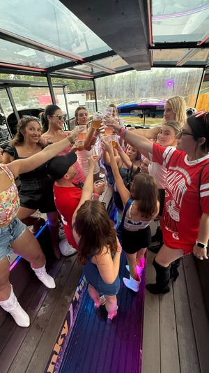 Group of friends on a covered outdoor party bus raising plastic cups in a toast under a glass roof with colorful LED lights, summer outfits and cowboy boots.