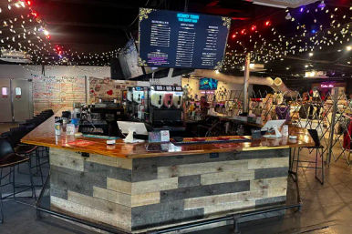 Rustic indoor bar island with wood-plank front and wooden countertop, POS stations and frozen-drink dispensers, overhead digital menu screen and twinkling string lights, high stools and open event space with stacked chairs in the background.