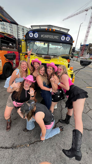 Cheerful group of women in pink cowboy hats and boots laughing and posing in front of a bright yellow party bus marked “#CARPOOLMIC,” parked on a city street with apartments and construction cranes in the background.