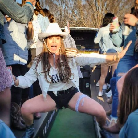 Energetic bride-to-be in a white cowboy hat and veil squats with arms out on a lively bachelorette party bus, wearing a 'BRIDE' top, black shorts and a garter, surrounded by friends holding drinks in an outdoor daytime setting.