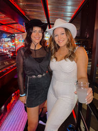 Two smiling women in cowboy hats posing on a neon-lit downtown bar balcony at night — one in a black dress, the other in a white outfit holding a drink.