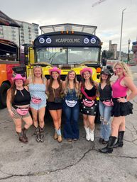 Seven women in cowgirl outfits and pink hats posing in front of a yellow party bus labeled "#CARPOOLMIC" parked on a downtown city street, wearing cowboy boots and pink fanny packs.