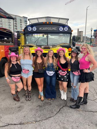 Seven women in cowgirl outfits and pink hats posing in front of a yellow party bus labeled "#CARPOOLMIC" parked on a downtown city street, wearing cowboy boots and pink fanny packs.