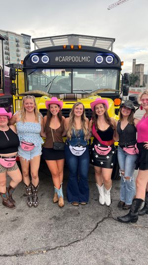 Seven friends in cowgirl outfits — pink hats, fanny packs and cowboy boots — posing in front of a yellow party bus parked on a city street during an outdoor event