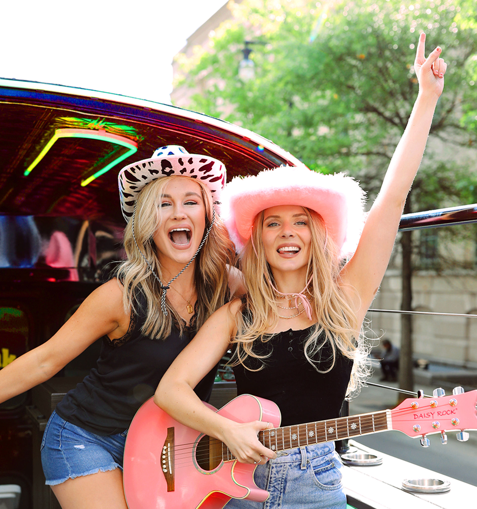 Two smiling young women wearing cowgirl hats—one in a pink fuzzy hat playing a pink acoustic guitar and the other in a cow-print hat—cheering from an open-top vehicle on a sunny city street.