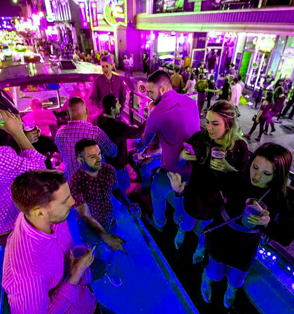 Nighttime downtown nightlife scene with young adults drinking and dancing on an illuminated rooftop party bus under neon signs and crowded sidewalks.