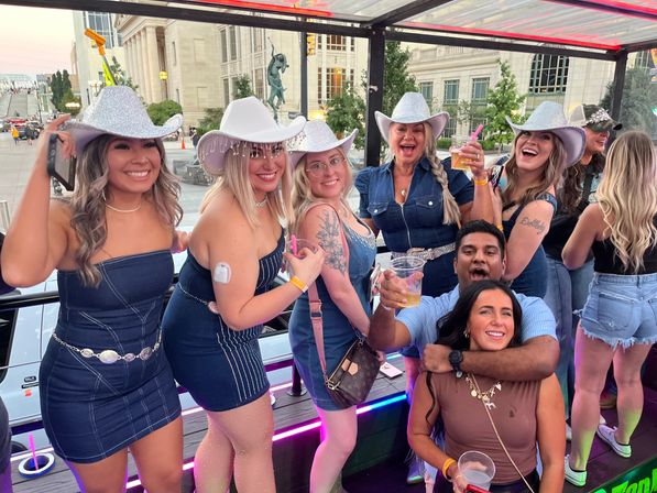 Smiling group in sparkly cowboy hats and denim outfits toasting drinks aboard an open-air party vehicle with a city street backdrop
