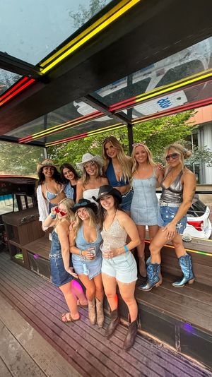 Smiling group of women in denim and cowgirl outfits posing on a neon-lit party bus with wooden benches and a city street visible through the windows, daytime celebration vibe