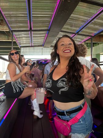 Smiling woman flashing a peace sign on a neon-lit party bus, wearing a black snake crop top, pink fanny pack and denim shorts, with friends in sunglasses and white boots laughing on a bench.