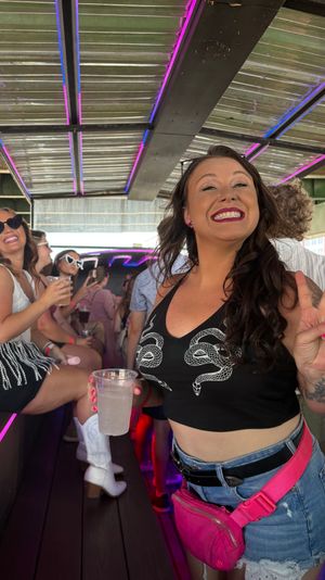 Neon-lit party bus interior with a smiling woman flashing a peace sign and holding a drink while friends in summer outfits laugh on bench seats.
