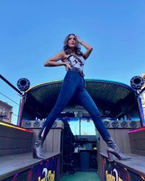 Low-angle shot of a woman striking a wide-legged pose on an open-air party bus in a city street, wearing a cow-print top, high-waist jeans and brown heeled ankle boots against a clear blue sky.
