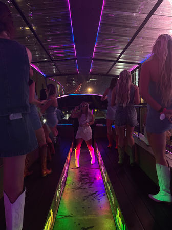 Neon-lit rooftop party bus at night — women in denim and cowboy boots dancing on a glowing LED walkway under pink and green lights