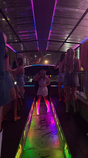 Neon-lit rooftop bar party at night with partygoers in denim and cowboy boots dancing on an illuminated runway, purple and green LED reflections on a wet floor and city lights in the background.
