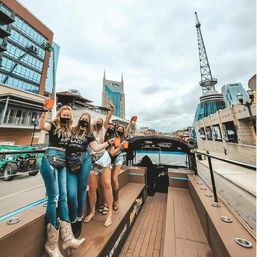 Group of masked women cheering with orange cups on an open-top party bus in downtown Nashville, with the iconic Batman Building and a tall radio tower in the cloudy skyline.