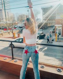 Young woman on a sunny downtown rooftop balcony raising her phone, wearing a sheer white crop top and jeans with red star patches, overlooking a city street with cars and pedestrians.