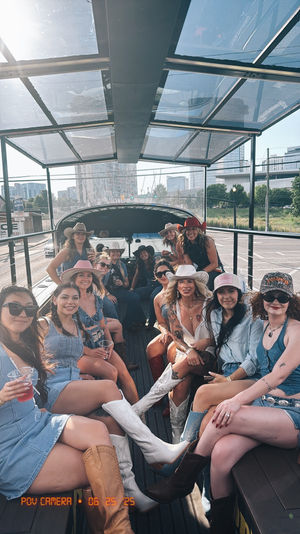 Group of smiling women in cowboy hats, denim and western boots sharing drinks on an open-air party vehicle with a downtown city skyline in the background.