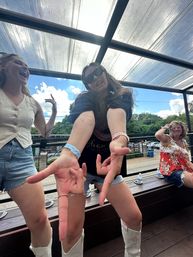 Three women having fun on a covered outdoor patio under a glass canopy on a sunny day, one wearing sunglasses reaching toward the camera with playful hand gestures while friends laugh on a bench