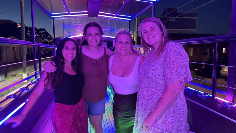 Four friends smiling on a neon-lit party bus at night, purple and blue LED lights, bachelorette-party vibe with city street in the background