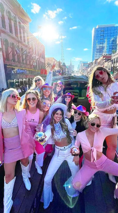 Group of women in pink and white cowgirl outfits and sunglasses throwing a lively bachelorette-style party on an open-top party bus down a sunny downtown avenue, holding sparkly drinks amid city buildings and traffic.