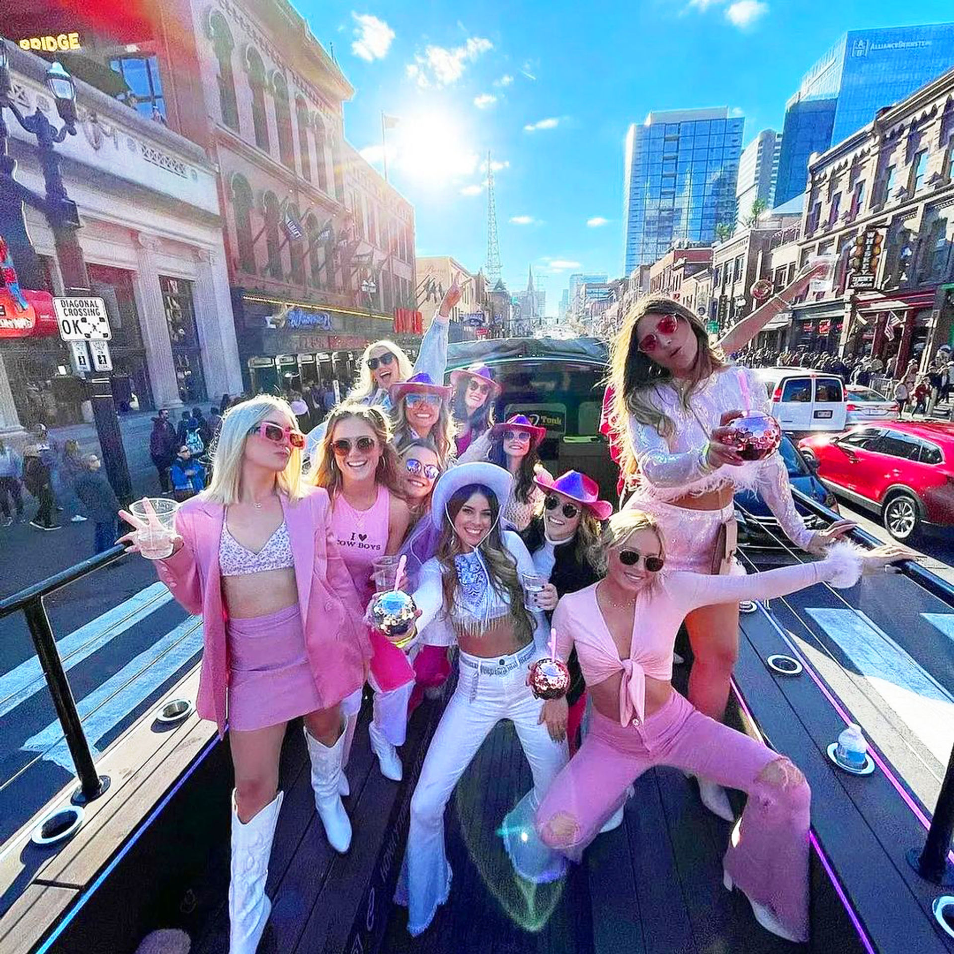 Group of women in pink and white cowgirl outfits and sunglasses throwing a lively bachelorette-style party on an open-top party bus down a sunny downtown avenue, holding sparkly drinks amid city buildings and traffic.