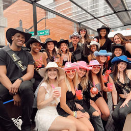 Smiling friends in cowboy hats and sashes raise colorful cocktails on a covered city rooftop patio during a bachelorette-style celebration.