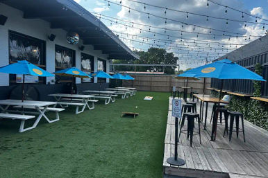 Outdoor patio seating with artificial turf, white picnic tables under blue umbrellas, cornhole boards and a raised wooden deck with high-top tables and string lights.