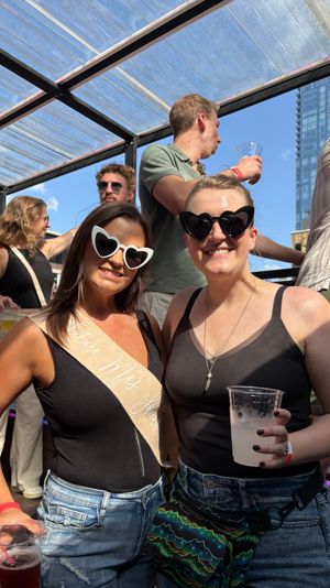 Two smiling women in heart-shaped sunglasses at a sunny urban rooftop party, one wearing a "Future Mrs." sash and both holding drinks with a city skyline and glass canopy overhead.