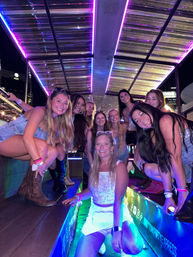 Smiling group of young women on a neon-lit party bus at night, wearing denim and cowboy boots beneath a colorful LED canopy with city lights in the background
