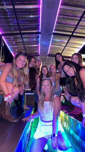 Neon-lit nighttime group shot of smiling friends posing inside a covered party trolley with purple and blue LED strips, city lights in the background, many wearing summer outfits and cowboy boots.