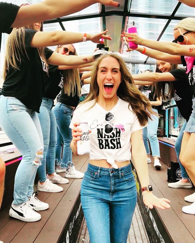 Excited woman holding a drink walks through cheering friends on a glass-roofed Nashville party deck, all in casual jeans and event tees with a bachelorette bash vibe.