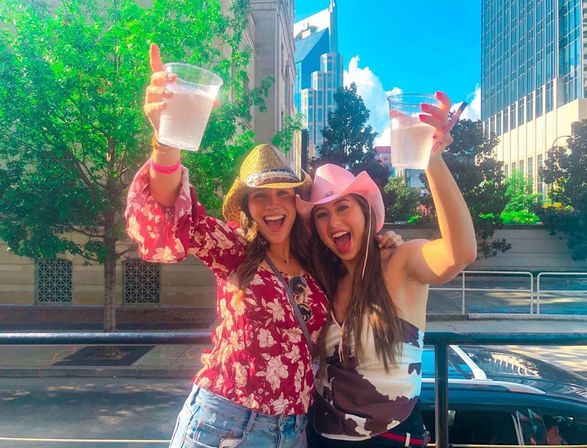 Two friends wearing cowgirl hats cheer with plastic cups at a sunny downtown street party, city skyline and trees in the background.