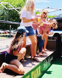 Four friends laughing on a sunny outdoor party deck by a parking lot, wearing summer outfits and sunglasses while holding cold drinks for a tailgate-style celebration.