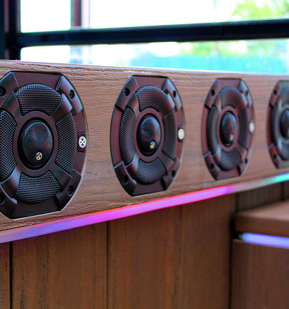 Close-up of four round black speakers mounted in a textured wooden panel beneath a window, with pink-purple LED accent lighting for a modern interior audio setup.
