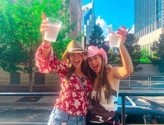 Two friends in straw and pink cowboy hats cheer with plastic cups on a sunny downtown city street, smiling in front of trees and tall office buildings.