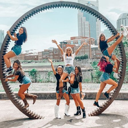 Bachelorette group of seven women posing on a giant circular gear sculpture by an urban riverfront, one lifted on friends’ shoulders while others climb the rim, wearing denim shorts and boots with a downtown skyline backdrop.