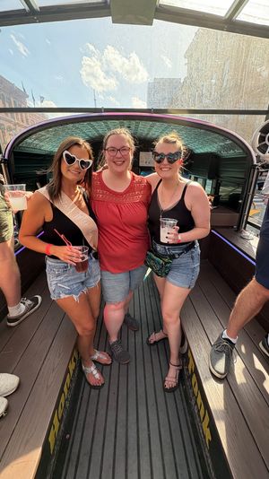 Three smiling women in sunglasses and summer shorts pose with drinks on a sunny downtown open-top party bike (pedal pub) with a glass canopy and city buildings in the background; one wears a bachelorette sash.