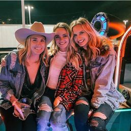 Three friends smiling in a neon-lit outdoor night scene — casual night out with denim jackets, ripped jeans and a cowboy hat, one holding a drink.