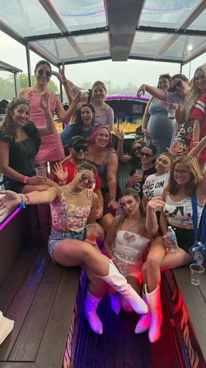 Group of friends partying on an open-air party bus with a clear roof and neon LED lights, wearing summer outfits and cowboy boots, holding drinks and smiling and waving.